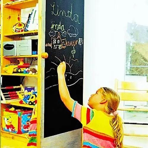 Child writing on a black chalkboard wall sticker with colorful chalk, showcasing creativity in a playful environment.