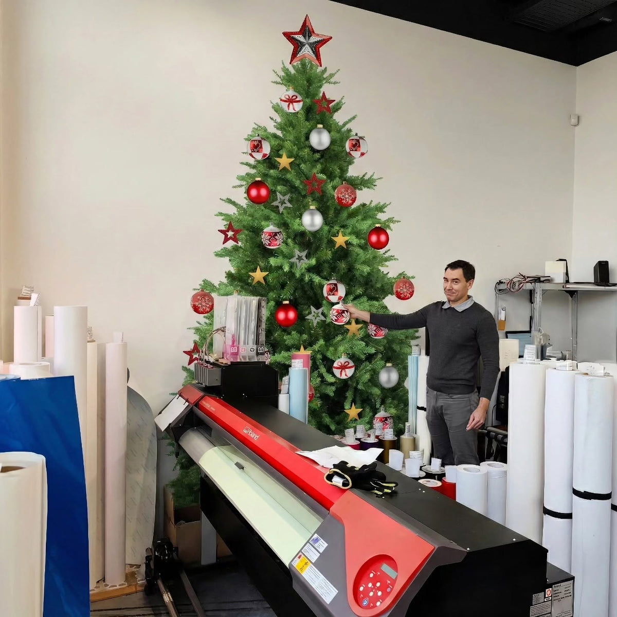 Christmas tree wall decal with separate ornaments on a workshop wall, beside a man, printer and rolls of material.