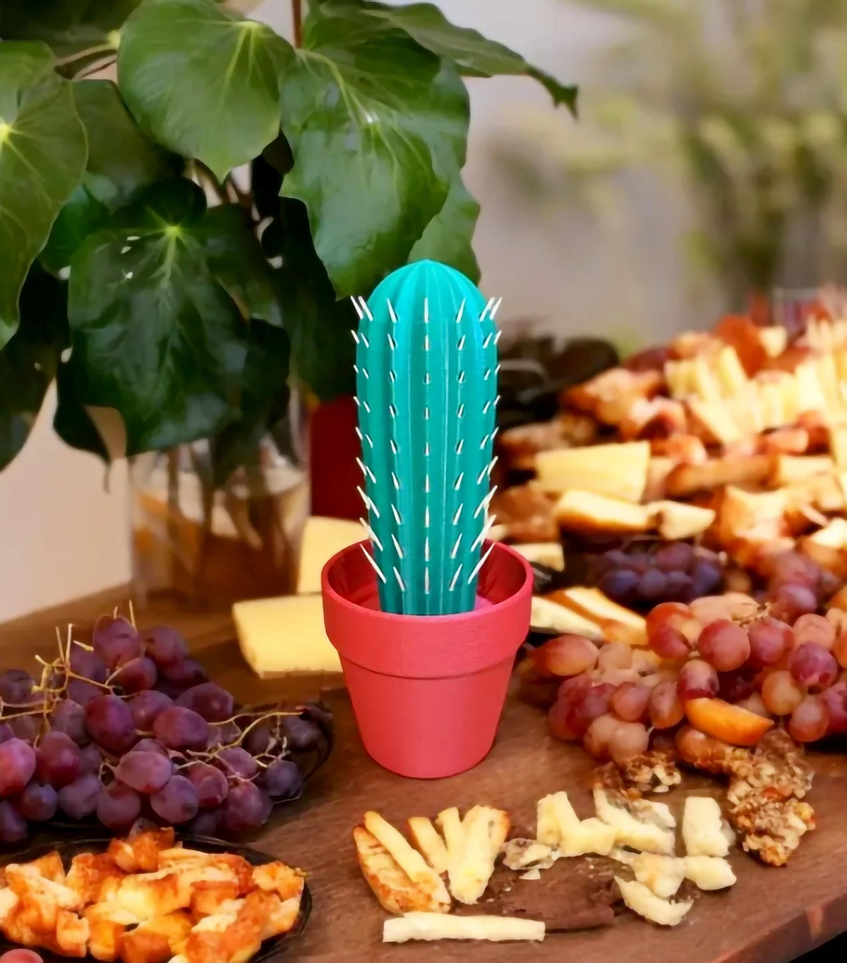 3D printed cactus toothpick holder in a pot, surrounded by fruit and cheese platters, adding fun decor to a party table.