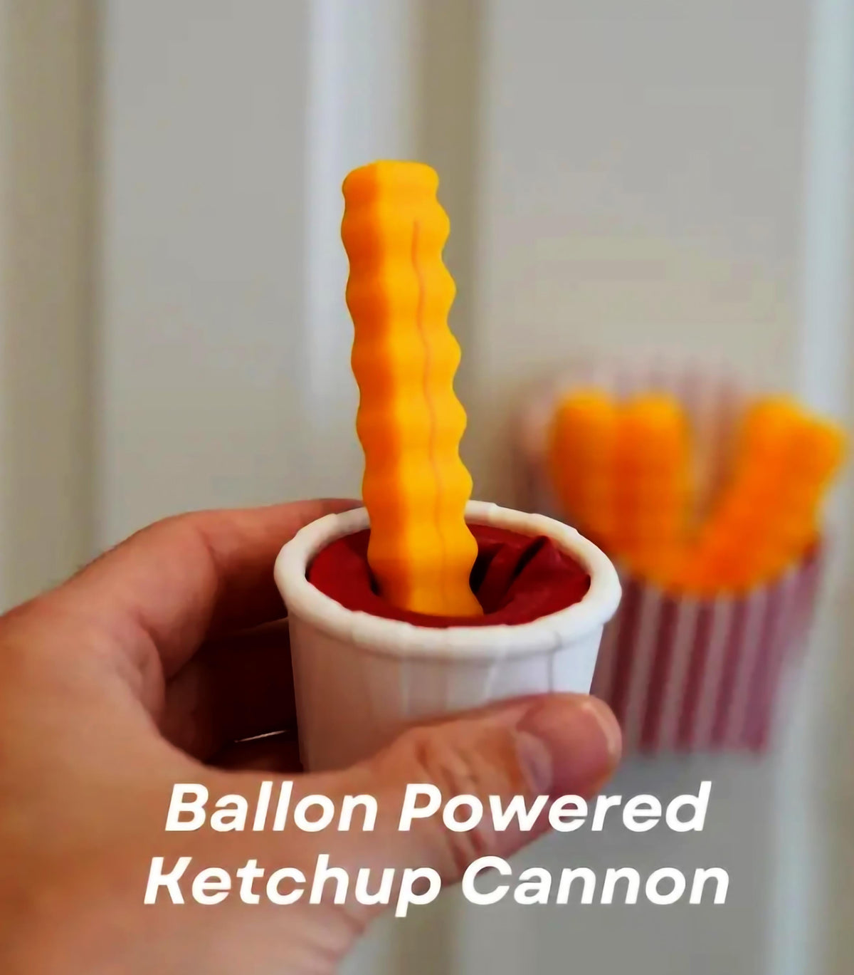 Hand holding a fun balloon-powered ketchup cannon in a cup, designed for playful kitchen snacks.