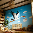 Flat illustration of a white stork with black-tipped wings flying in a bright blue sky with clouds.