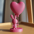 3D printed rabbit figurine on a circular base holding a heart-shaped balloon, photographed on a wooden surface.