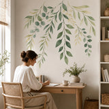 Woman sitting at a wooden desk beneath cascading green leaf wall stickers in a bright room.