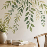 Close-up of hanging green botanical wall stickers above a wooden desk with a white vase and books.