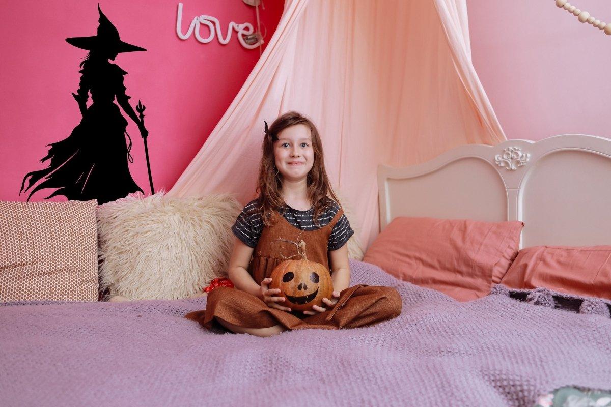 Young girl sitting on a bed with a pumpkin, featuring a witch silhouette decal on the wall for Halloween decor.