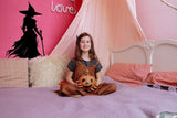 Young girl sitting on a bed with a pumpkin, featuring a witch silhouette decal on the wall for Halloween decor.