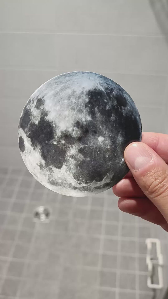 Person holding a circular moon decal sticker against a tiled background, showcasing its glow-in-the-dark design.