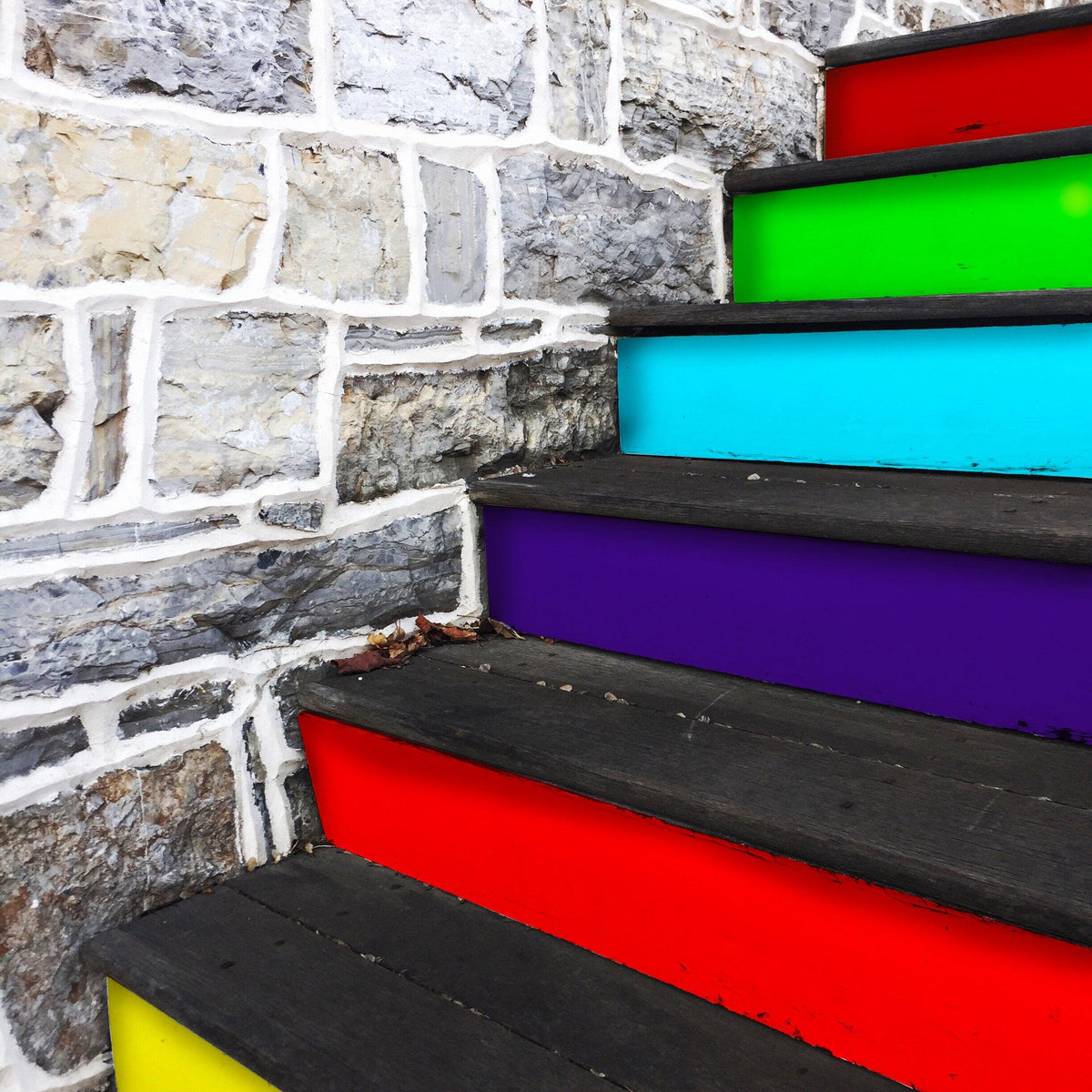 Vibrant rainbow self-adhesive stairs risers stickers on wooden staircase against a stone wall.