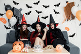 Three women in witch costumes celebrating Halloween with bats decorations and treats on a festive table.