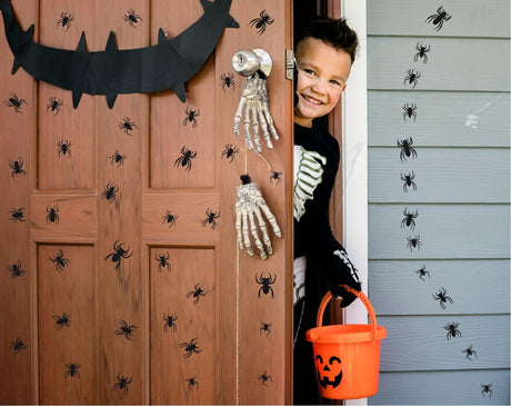 Child in skeleton costume smiling by Halloween spider wall decals on door, holding pumpkin candy bucket.