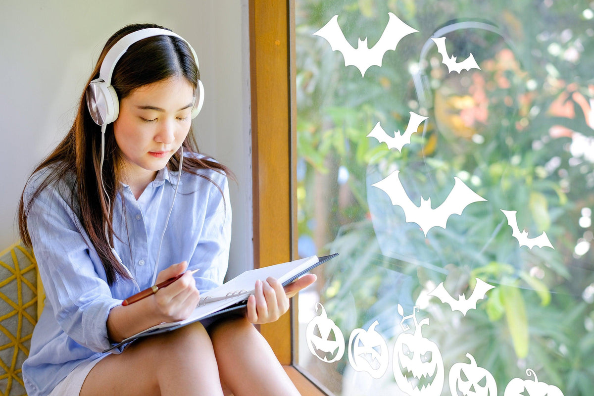 Girl wearing headphones sketching by a window with Halloween window decals of bats and pumpkins.